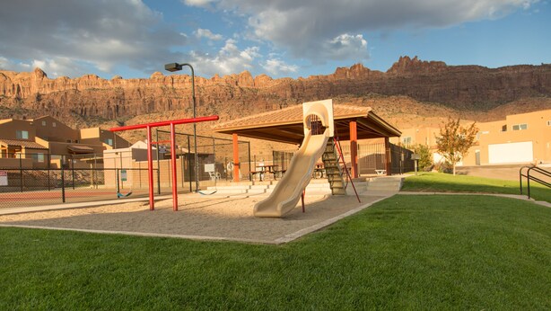 A playground with a slide and swings, surrounded by a fenced area, with buildings and a mountain in the background. - Best Rental in Town. (Moab)