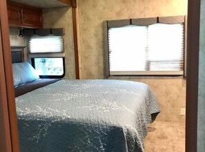 A compact RV bedroom with a bed, a window with blinds, and a small kitchenette area.