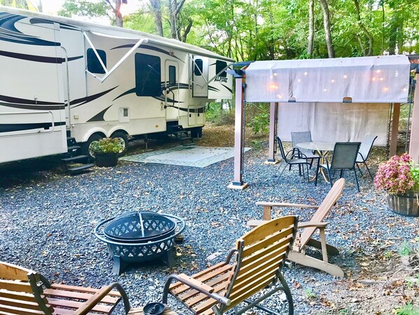 A recreational vehicle (RV) parked in a gravel area with a covered outdoor seating area, a fire pit, and a table with chairs.
