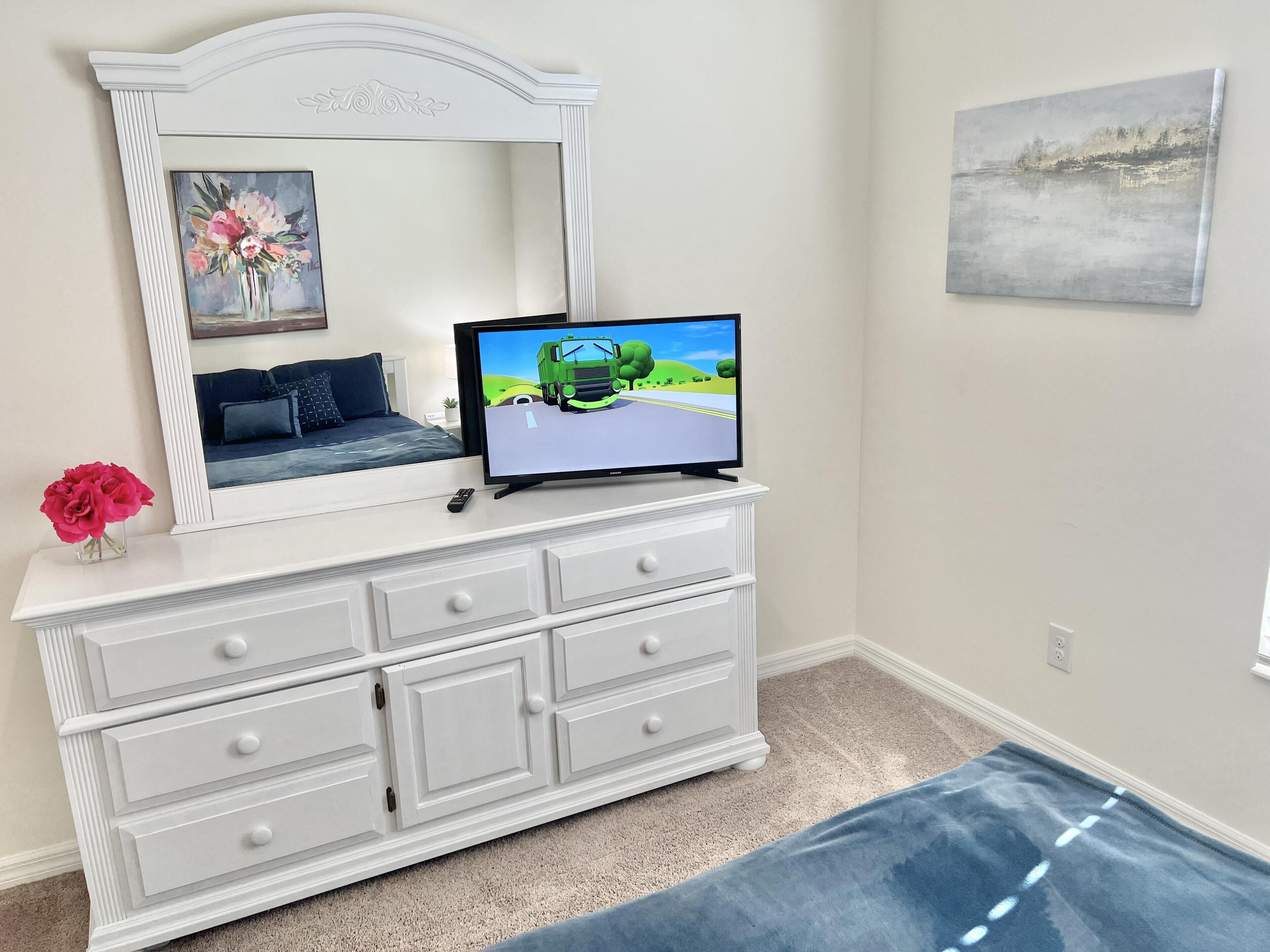 A white dresser with a mirror, a television displaying a landscape, and a vase of pink flowers.