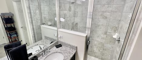 A modern bathroom with a granite countertop sink, a large mirror, and a glass shower enclosure.