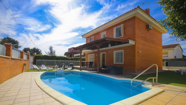 Une piscine avec une terrasse carrelée, une maison en briques dotée d’un patio couvert et un espace clôturé avec des chaises longues.