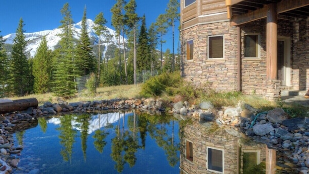 A lodge with a stone exterior, a wooden deck, and a clear pond reflecting the surrounding trees and mountains.