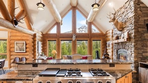 A log cabin interior with a stone fireplace, large windows, and a kitchen with Jenn-Air appliances.