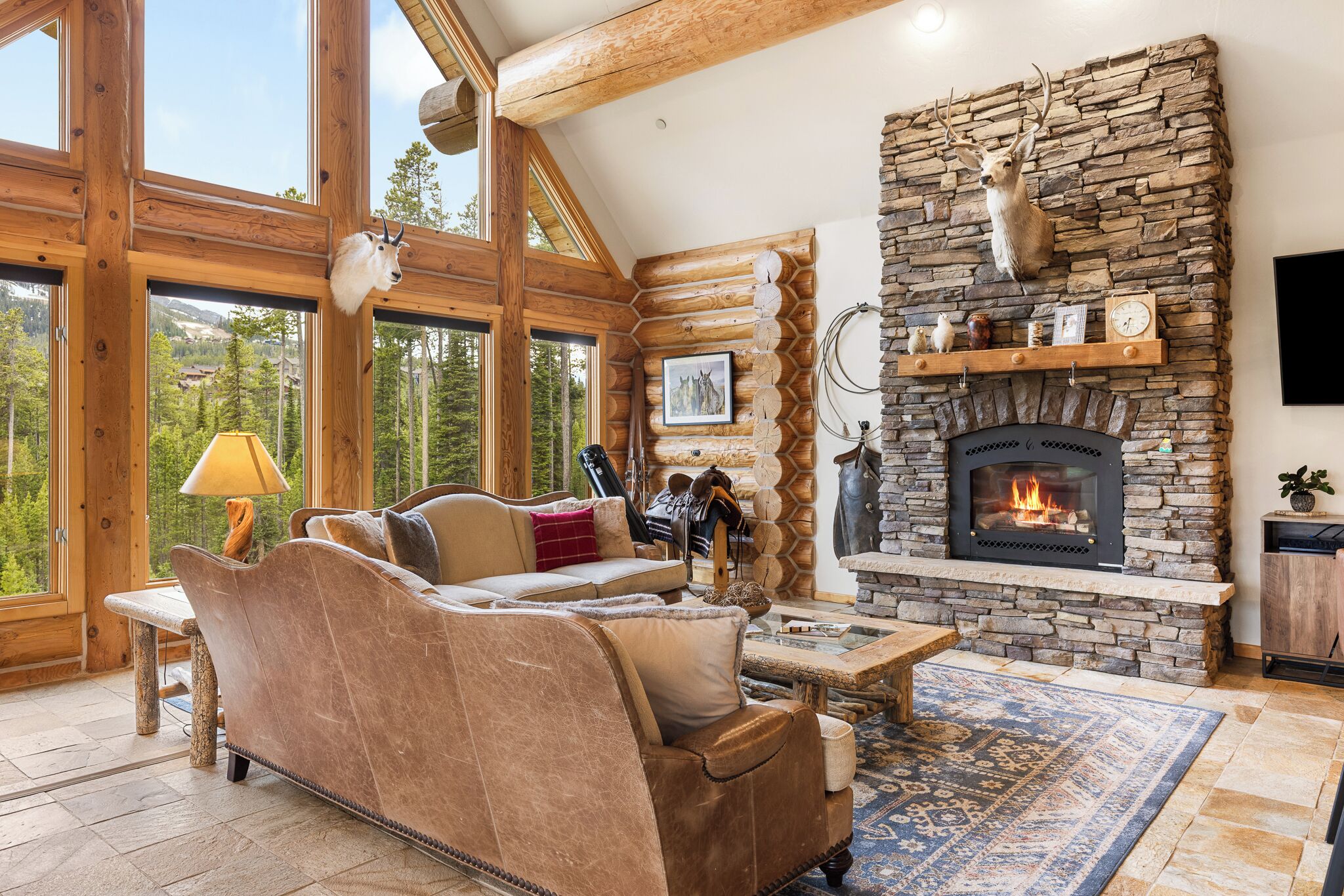 A log cabin interior with a stone fireplace, leather sofa, and large windows overlooking a forest.