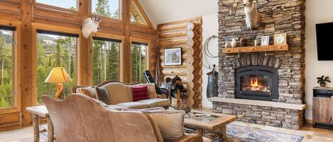 A log cabin interior with a stone fireplace, leather sofa, and large windows overlooking a forest.