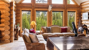 A log cabin interior with a large window, wooden walls, a sofa, a table, and a mounted animal head.