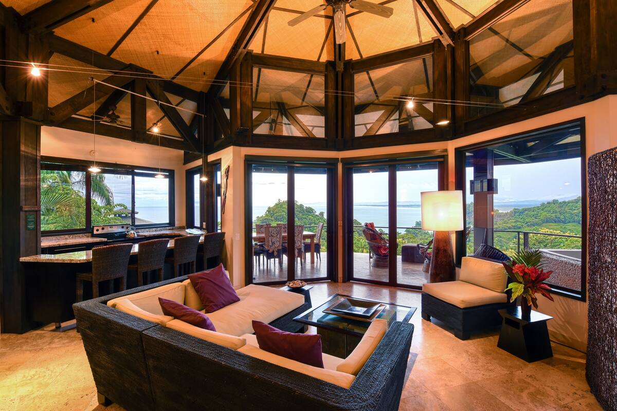 A spacious living area with a large sofa, a coffee table, and a dining area with chairs and a table. There are large windows offering a view of the outdoors.