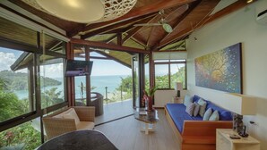 Une chambre moderne dotée d’une grande fenêtre offrant une vue sur la mer et une végétation luxuriante. On y trouve un canapé, une table et un téléviseur fixé au mur.