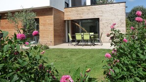 Une maison moderne avec une façade en bois, une porte d’entrée en verre et un patio agrémenté de chaises vertes.