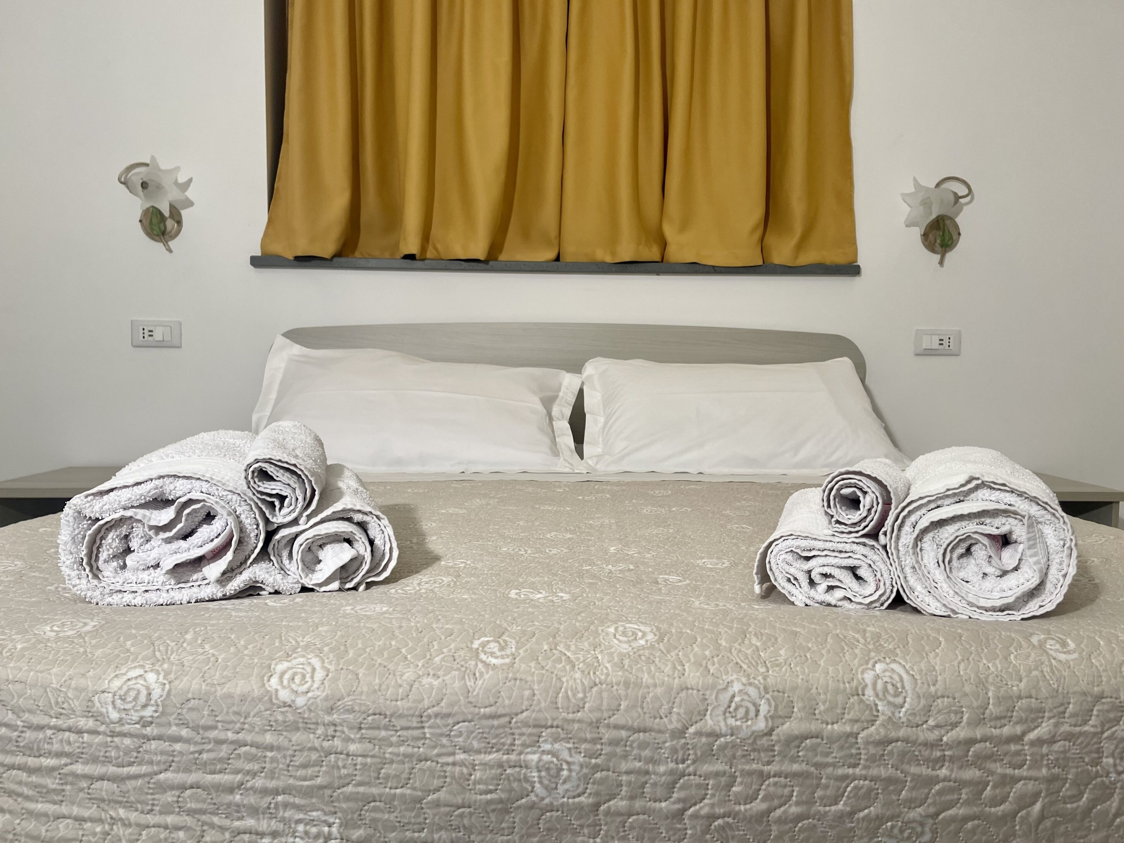 Een opgemaakt bed met opgerolde handdoeken, een bed met dessin en een geel gordijn.