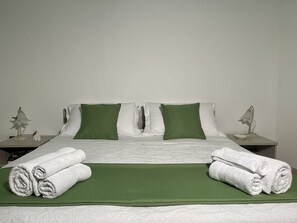Een keurig opgemaakt bed met groen-wit beddengoed, twee nachtkastjes met lampen en opgerolde witte handdoeken op het bed.