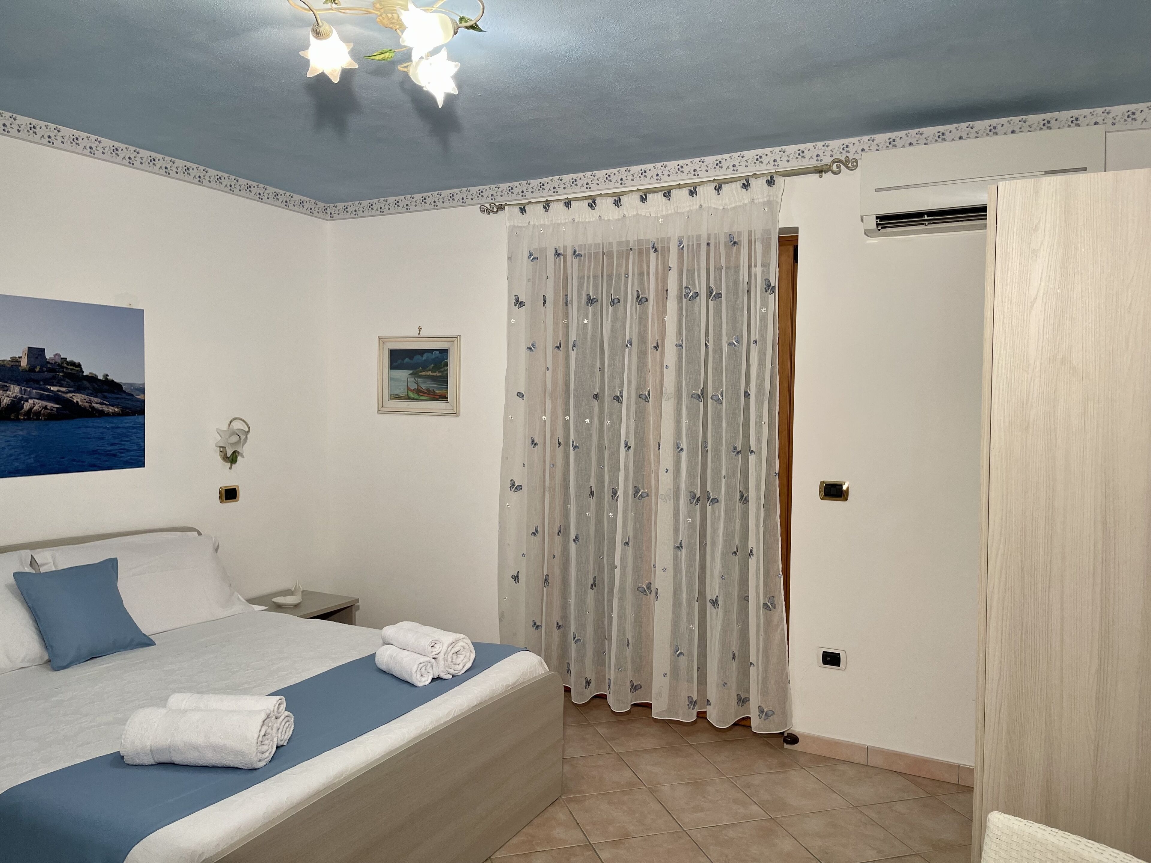 Una cama bien tendida con ropa de cama azul y almohadas blancas, una mesita de noche con una lámpara, un cuadro en la pared y una ventana con cortina.