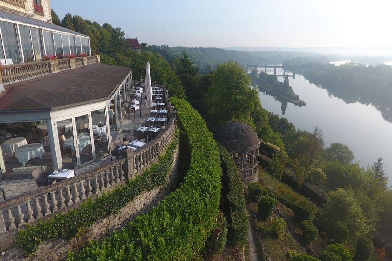 A riverside restaurant with outdoor seating overlooking a calm river and a bridge in the distance.