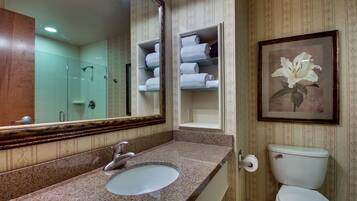 A bathroom with a granite countertop sink, a large mirror, a framed floral artwork, and a toilet with a towel shelf above it.
