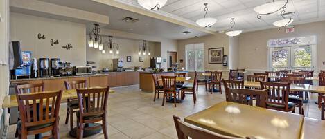 A spacious dining area with wooden tables and chairs, a kitchen area with coffee machines, and a large window providing natural light.