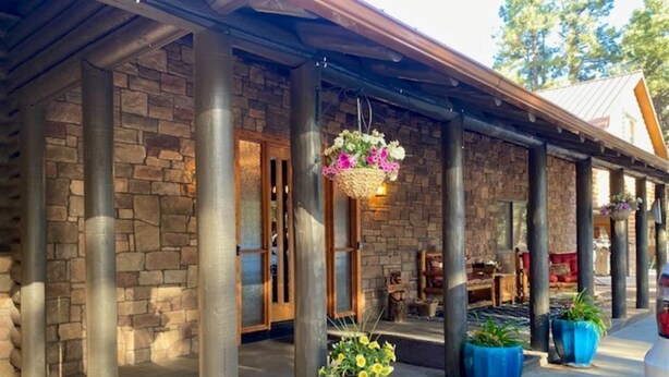 A stone building with a covered porch, columns, hanging flower baskets, and potted plants. - RAIN VALLEY RETREAT on 6 STUNNING acres! GAMES/GRILL/AC! (Flagstaff)
