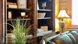 A cozy living room with a leather sofa, wooden shelves with books and decorative items, a potted plant, and a stone wall. - RAIN VALLEY RETREAT on 6 STUNNING acres! GAMES/GRILL/AC! (Flagstaff)