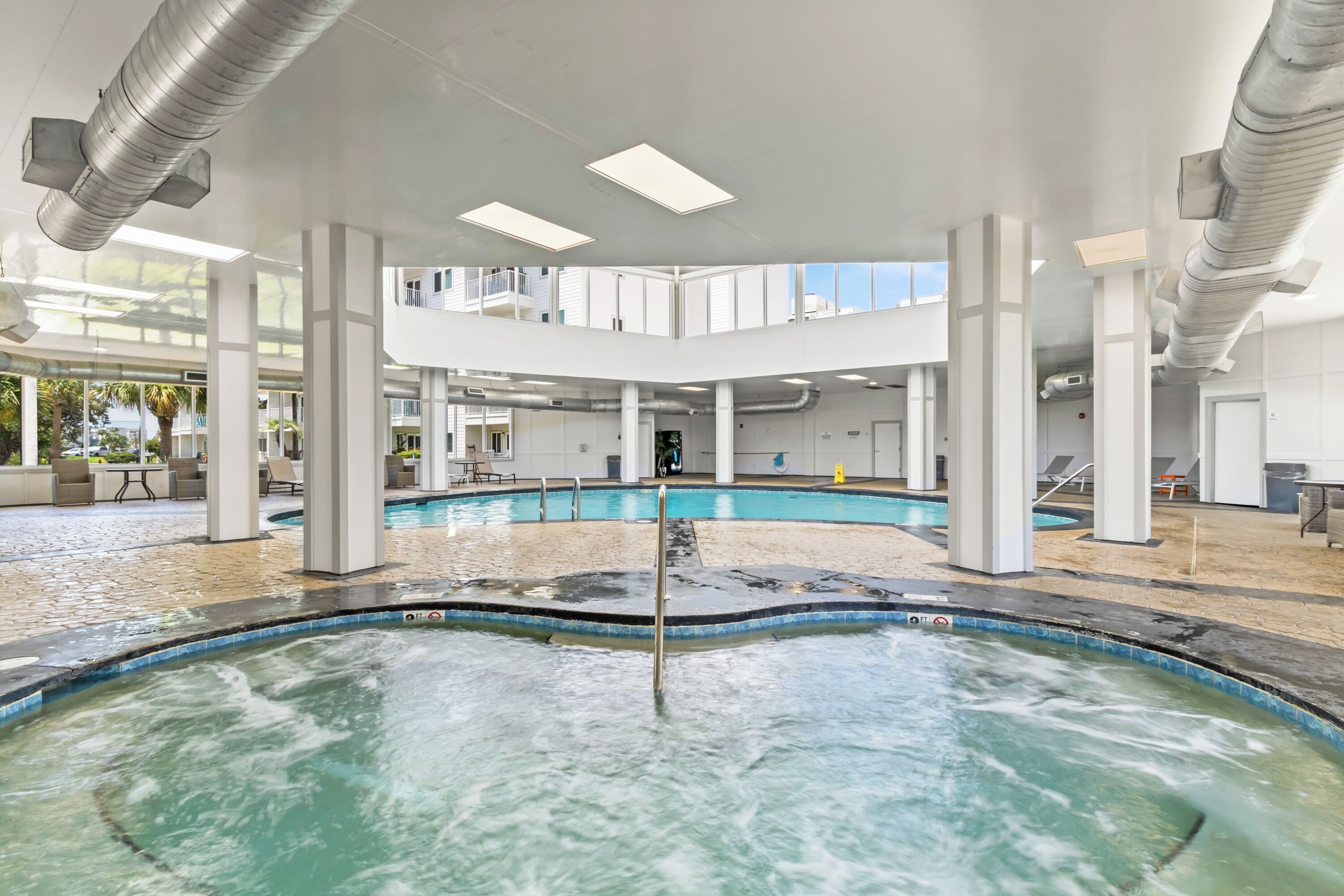 A modern indoor pool area with multiple pools, white pillars, and a clear view of the outside.