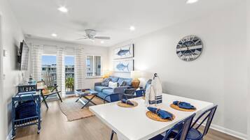 A spacious living area with a white ceiling fan, a dining table with blue chairs, and a large wall clock.