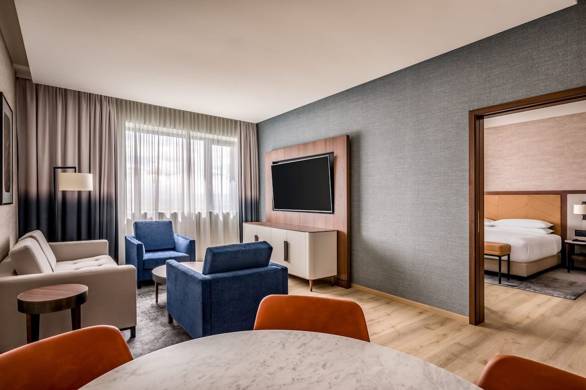 Ein modernes Hotelzimmer mit flachbildfernseher an der Wand, einer Couch, einem blauen Sessel, einem kleinen runden Tisch und einem Bett mit Holz-Kopfteil.