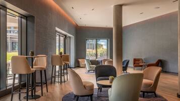 A modern lounge area with multiple seating options, including chairs and tables, arranged around a central column. Large windows provide natural light and views of the outdoors.