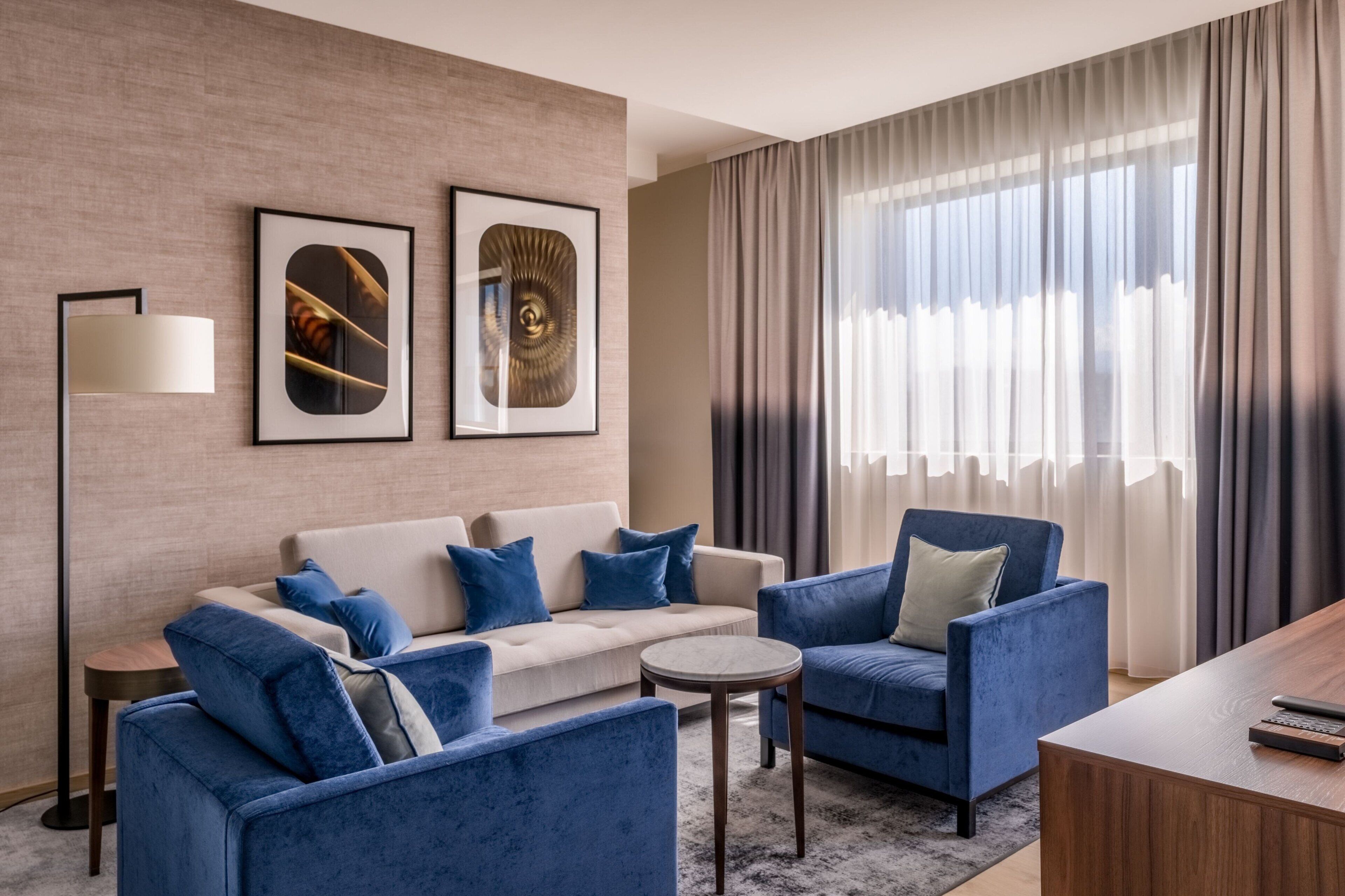 A modern hotel room with a beige sofa, blue armchairs, a round coffee table, and a wooden desk. There are framed artworks on the wall and a large window with curtains.