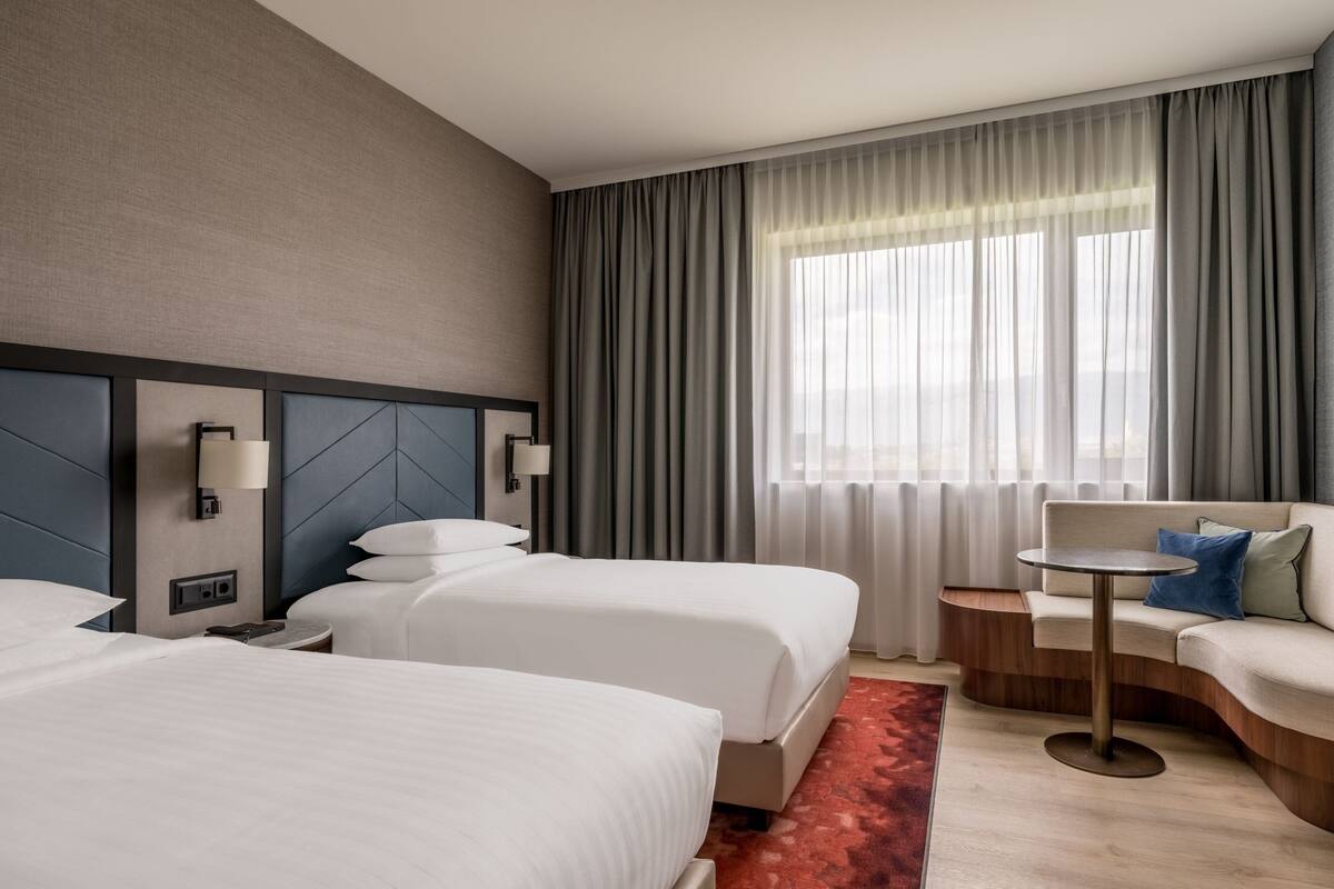 Une chambre d’hôtel avec deux lits, un canapé, une petite table et une vue sur la ville grâce à de grandes fenêtres.