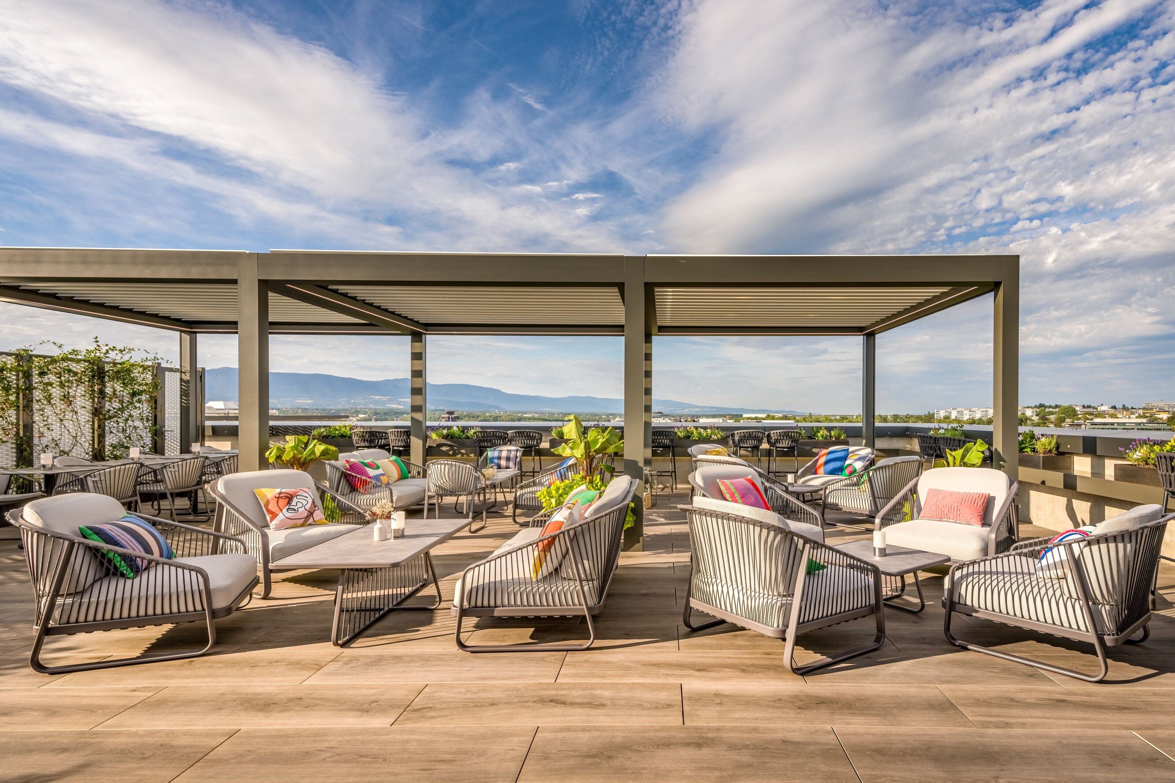 Ein Außenbereich mit Metallstühlen und -tischen, umgeben von einer Pergola, mit Blick auf eine malerische Berglandschaft und ein Gewässer.