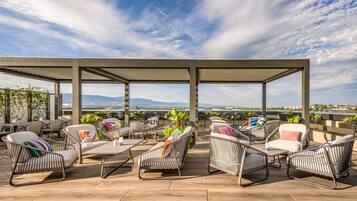 Ein Außenbereich mit Metallstühlen und -tischen, umgeben von einer Pergola, mit Blick auf eine malerische Berglandschaft und ein Gewässer.