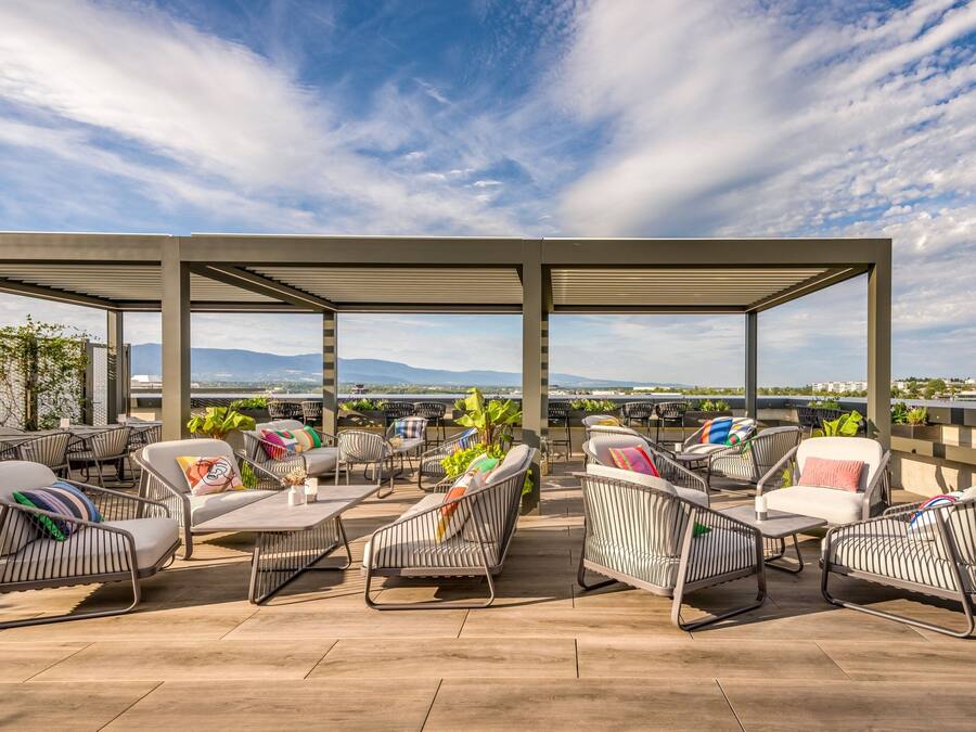 Ein Außenbereich mit Metallstühlen und -tischen, umgeben von einer Pergola, mit Blick auf eine malerische Berglandschaft und ein Gewässer.