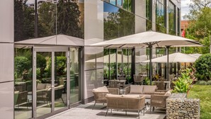 Un patio al aire libre con mobiliario de mimbre y sombrillas, amplias ventanas de vidrio y una jardinera de piedra.