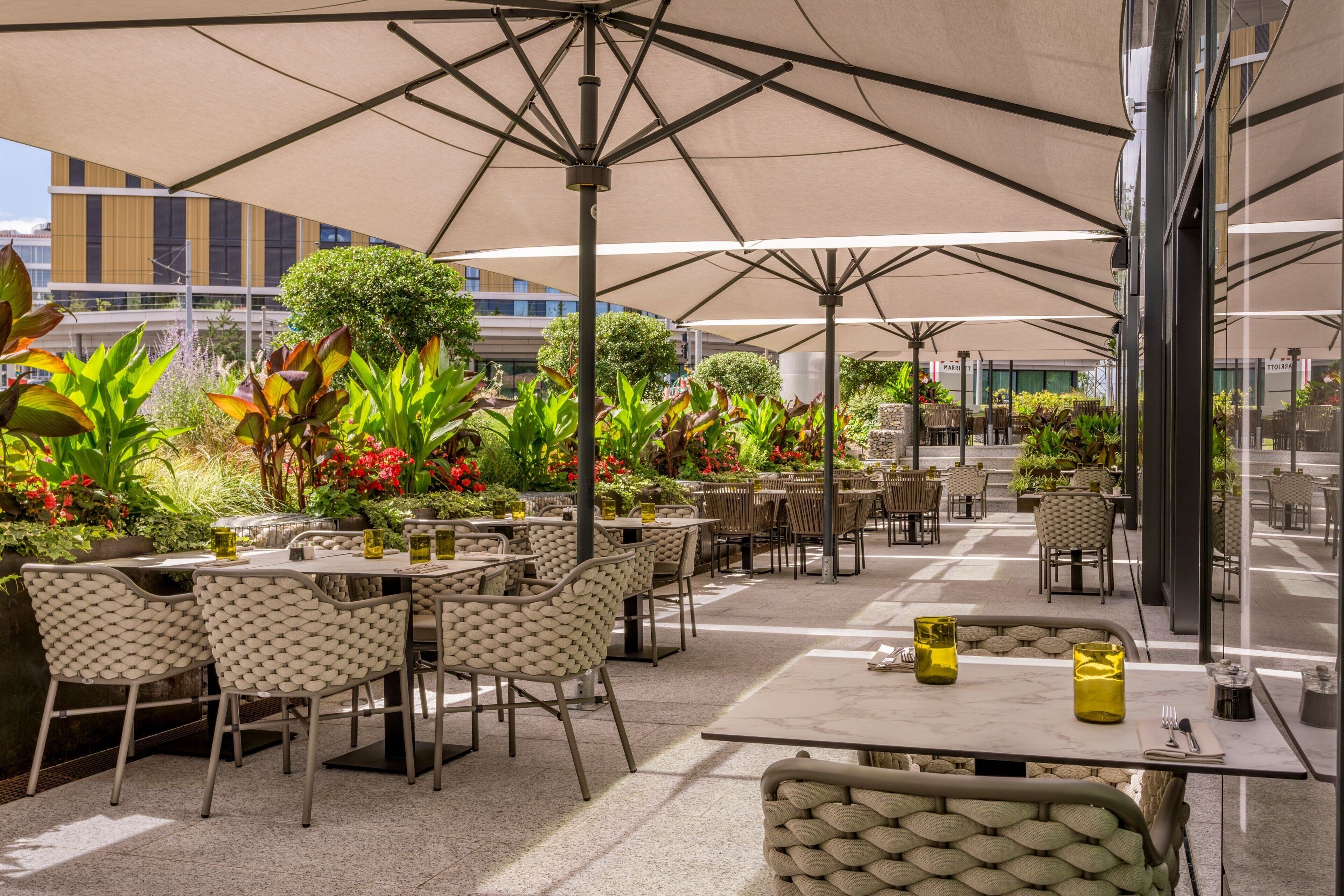 Café al aire libre con mesas y sillas, rodeado de plantas y sombrillas.
