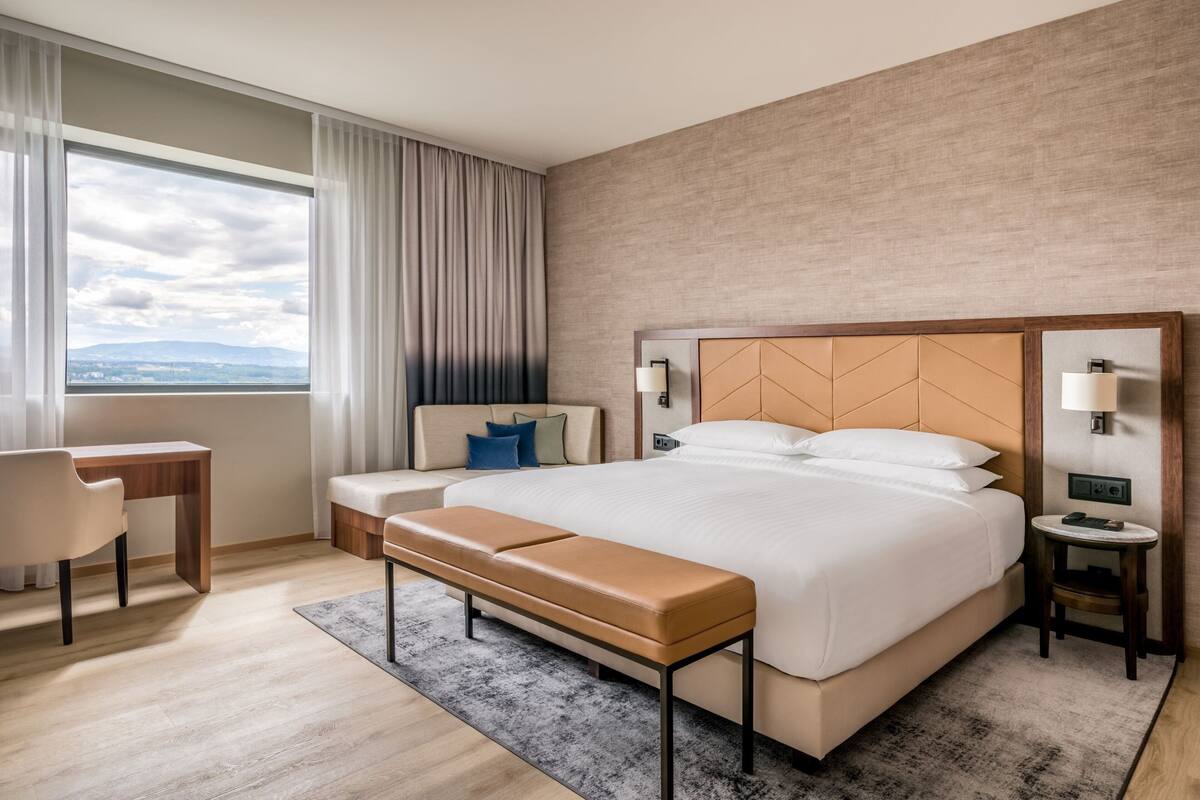Une chambre d’hôtel moderne, dotée d’un grand lit, d’un bureau, d’une banquette et offrant une vue sur l’extérieur.
