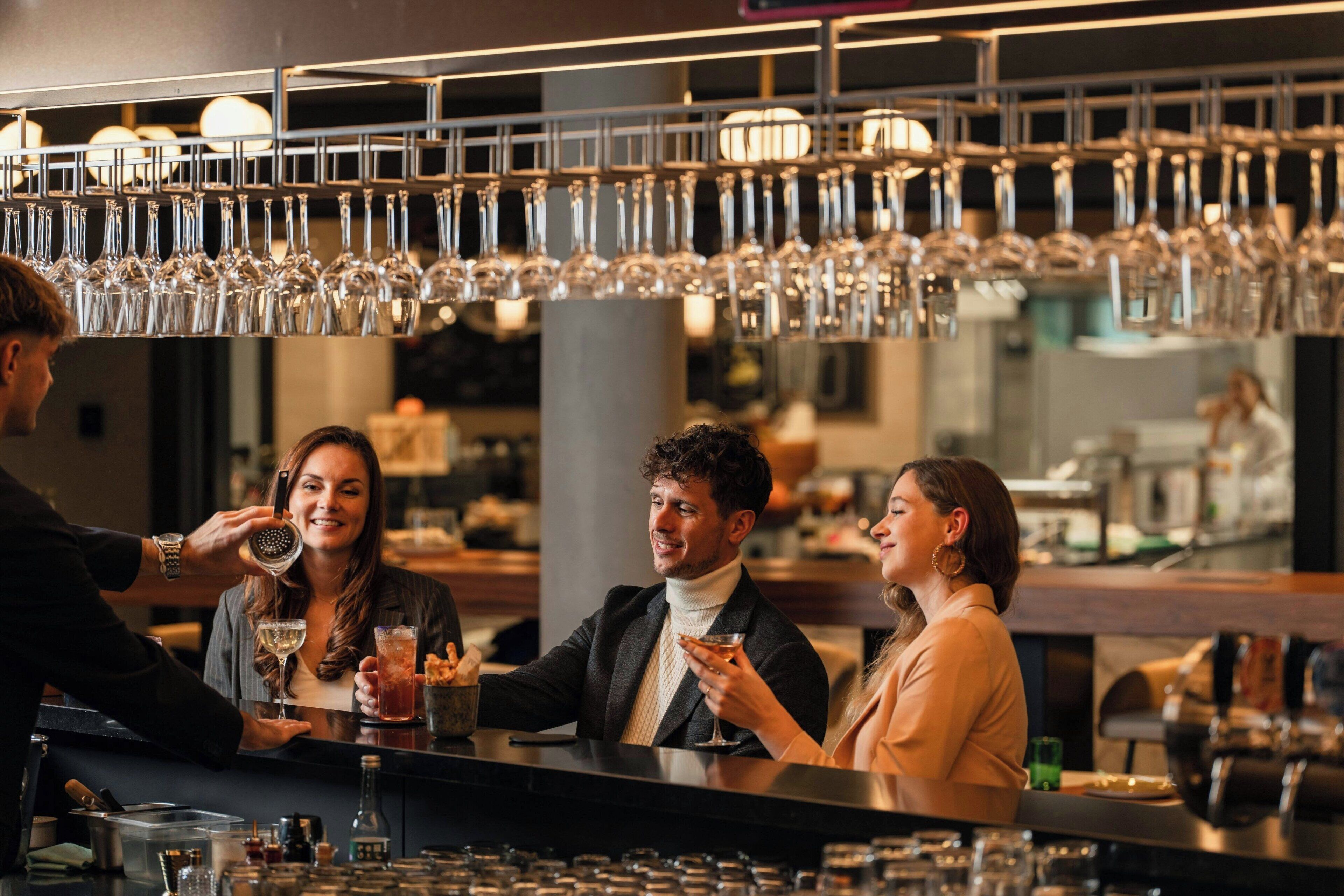 Een barruimte met verschillende hangende glazen, een barman en gasten die aan de bar zitten.