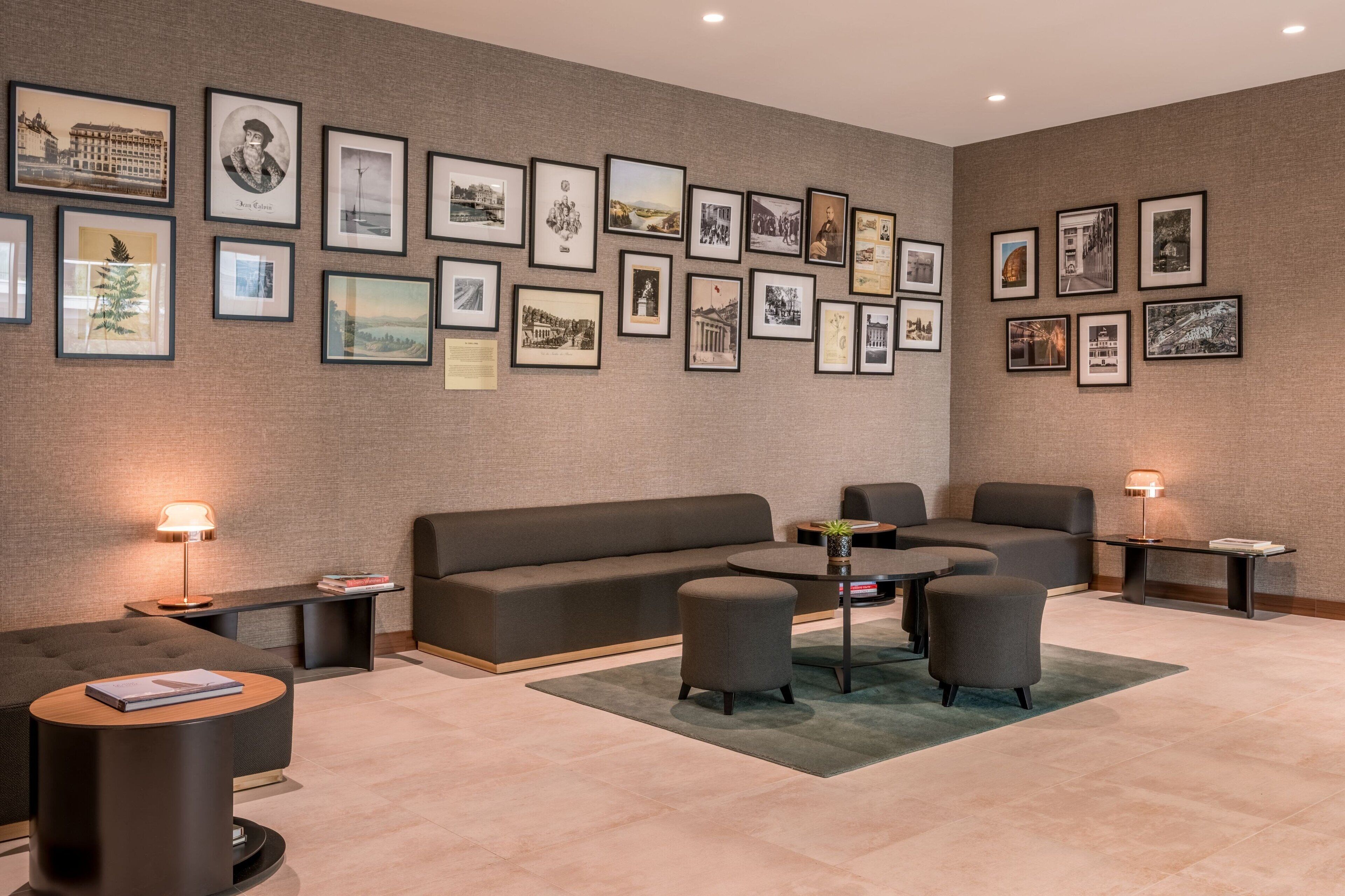 A modern lounge area with a sofa, armchairs, and a coffee table. There are framed pictures on the wall and two lamps on side tables.