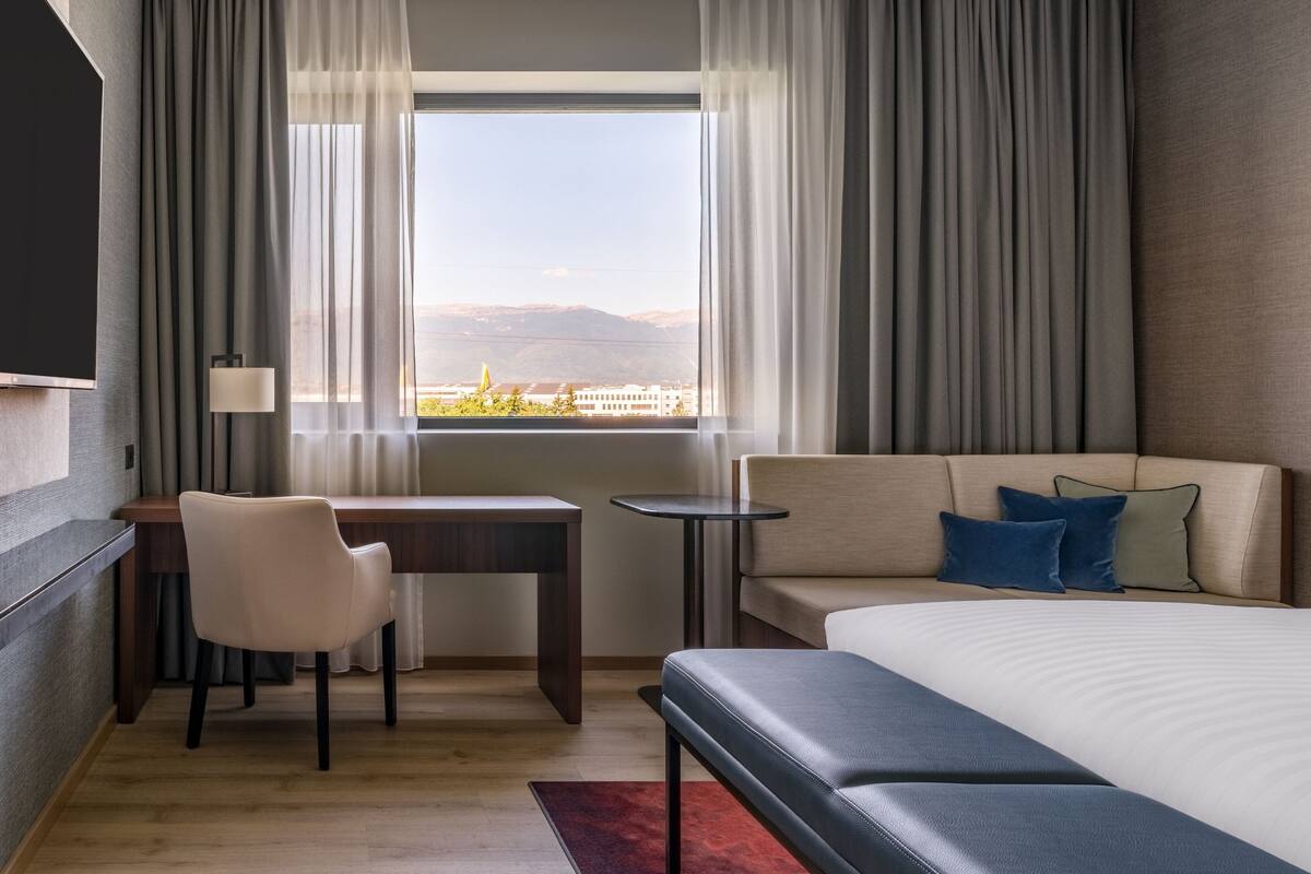 Une chambre d’hôtel avec un lit, un bureau, une chaise, un canapé et une vue sur les montagnes par la fenêtre.