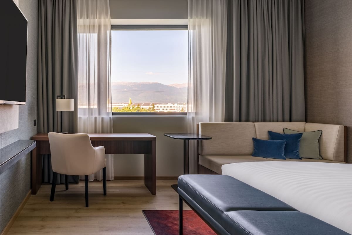 Ein Hotelzimmer mit Bett, Schreibtisch, Sessel und einer Aussicht auf Berge durch das Fenster.