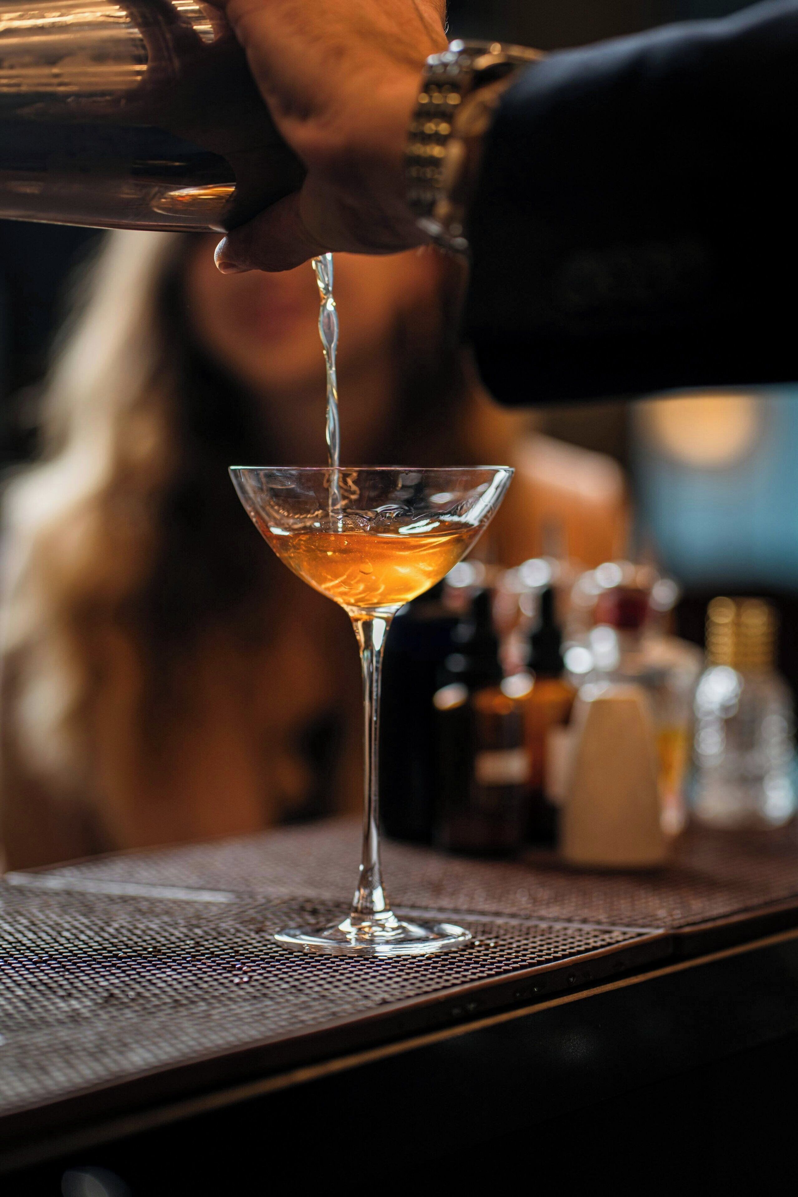 Un verre à martini posé sur le comptoir d’un bar, et une main y verse une boisson.