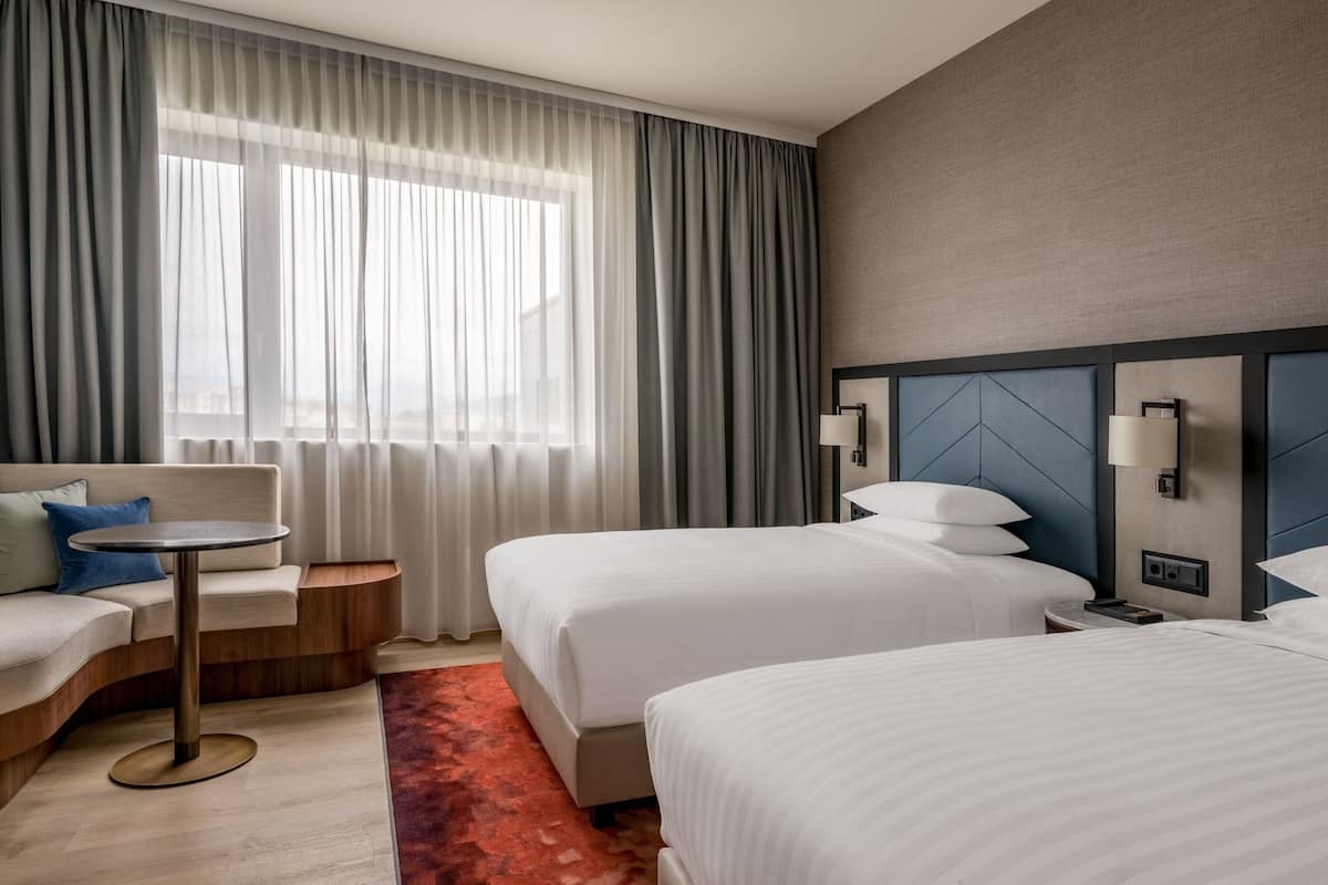 Une chambre d’hôtel avec deux lits, un canapé, une petite table et une vue sur la ville grâce à de grandes fenêtres.