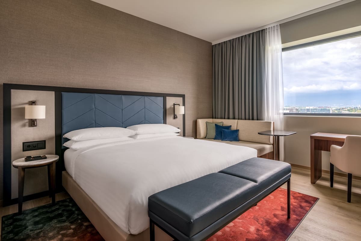 Ein Hotelzimmer mit einem großen Bett, einer Bank, einem Schreibtisch und Blick auf die Stadt.