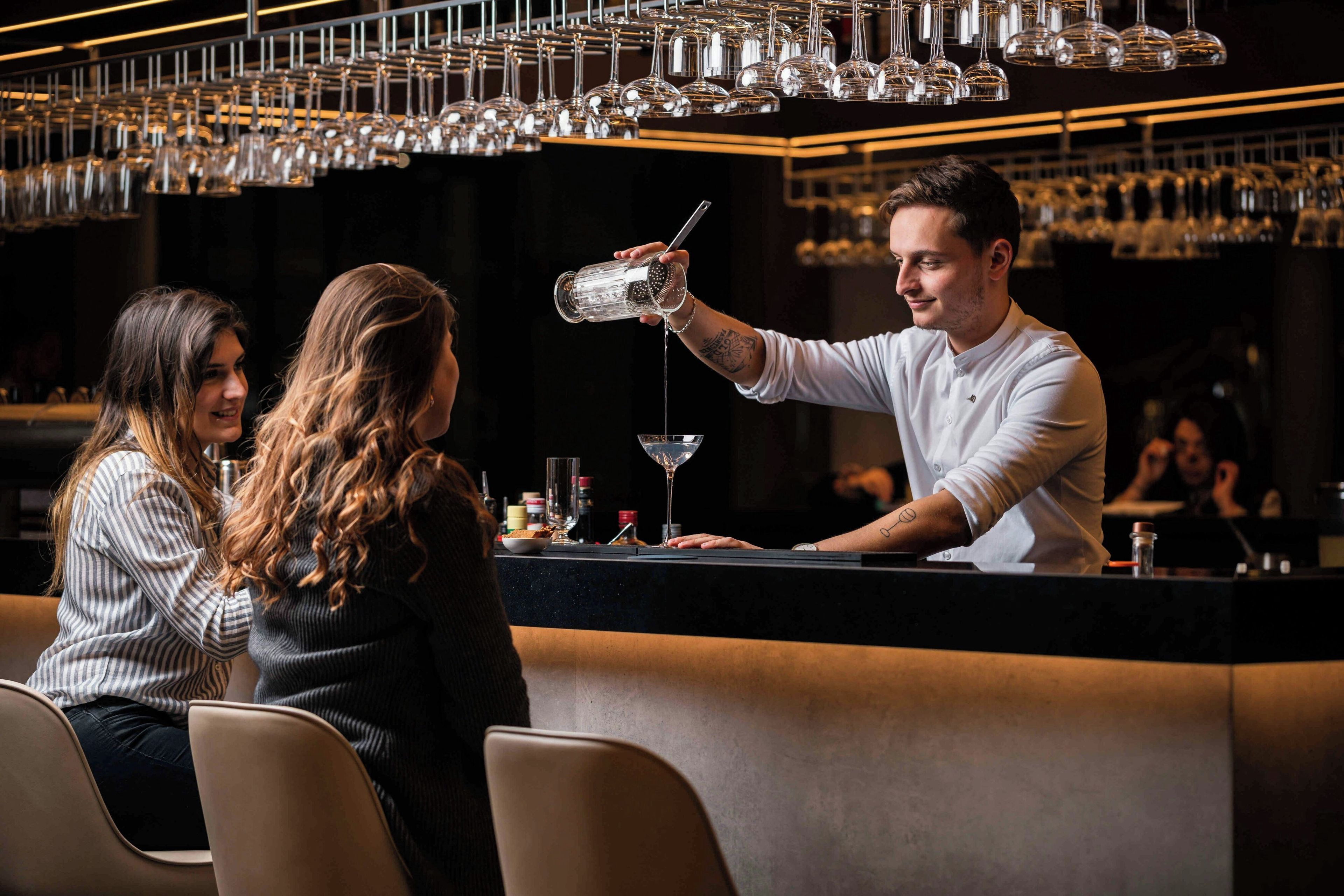 Eine Bar mit einem Barkeeper, der ein Getränk einschenkt, einer Theke mit verschiedenen Bartools und einer Wand voller hängender Weingläser.