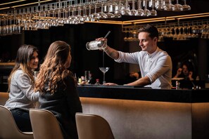 A bar setting with a bartender pouring a drink, a counter with various bar tools, and a backdrop of hanging wine glasses. - Geneva Marriott Hotel (Meyrin)
