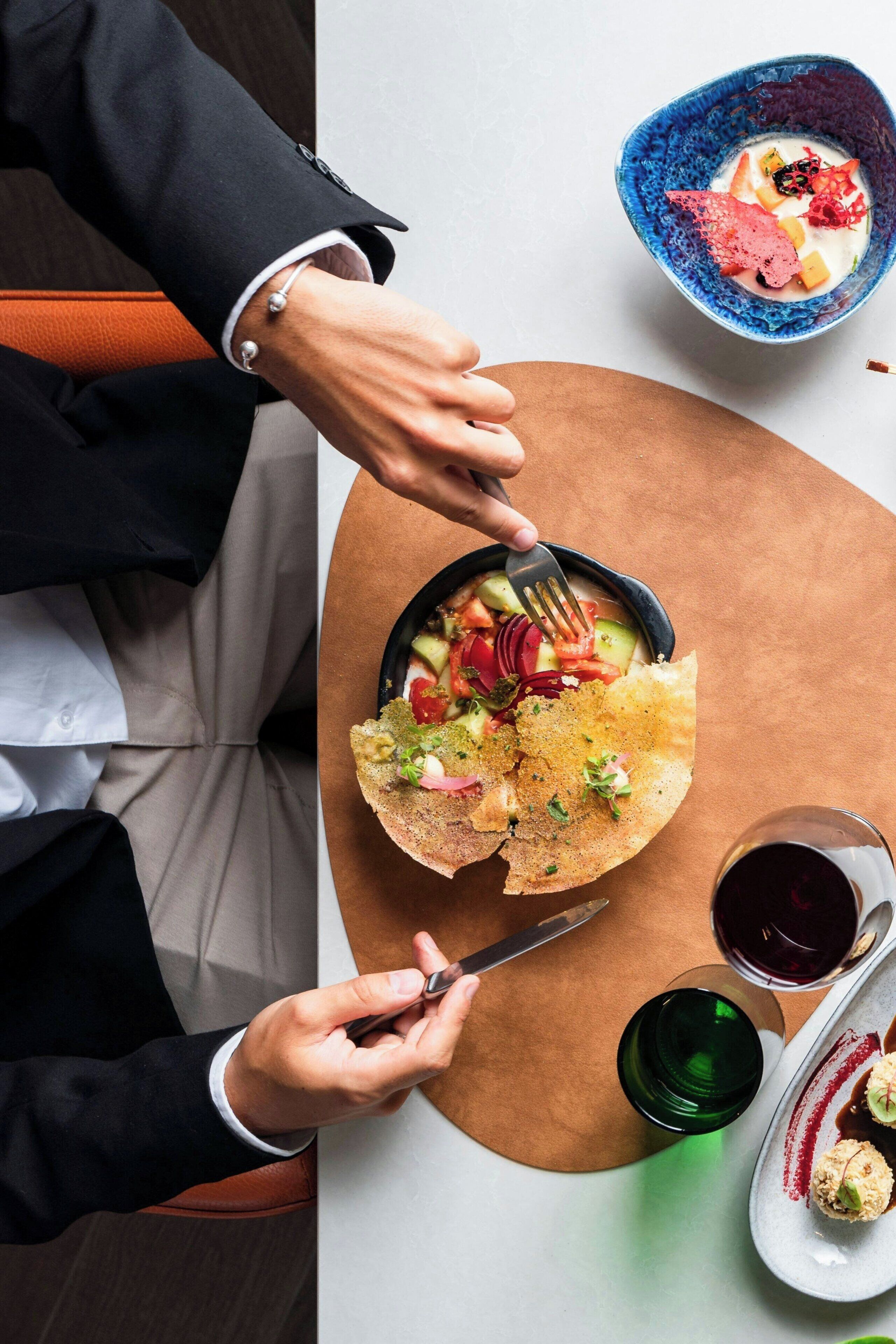 Uma pessoa de terno preto jantando à mesa, com uma salada numa tigela, um prato de comida e um copo de vinho tinto.
