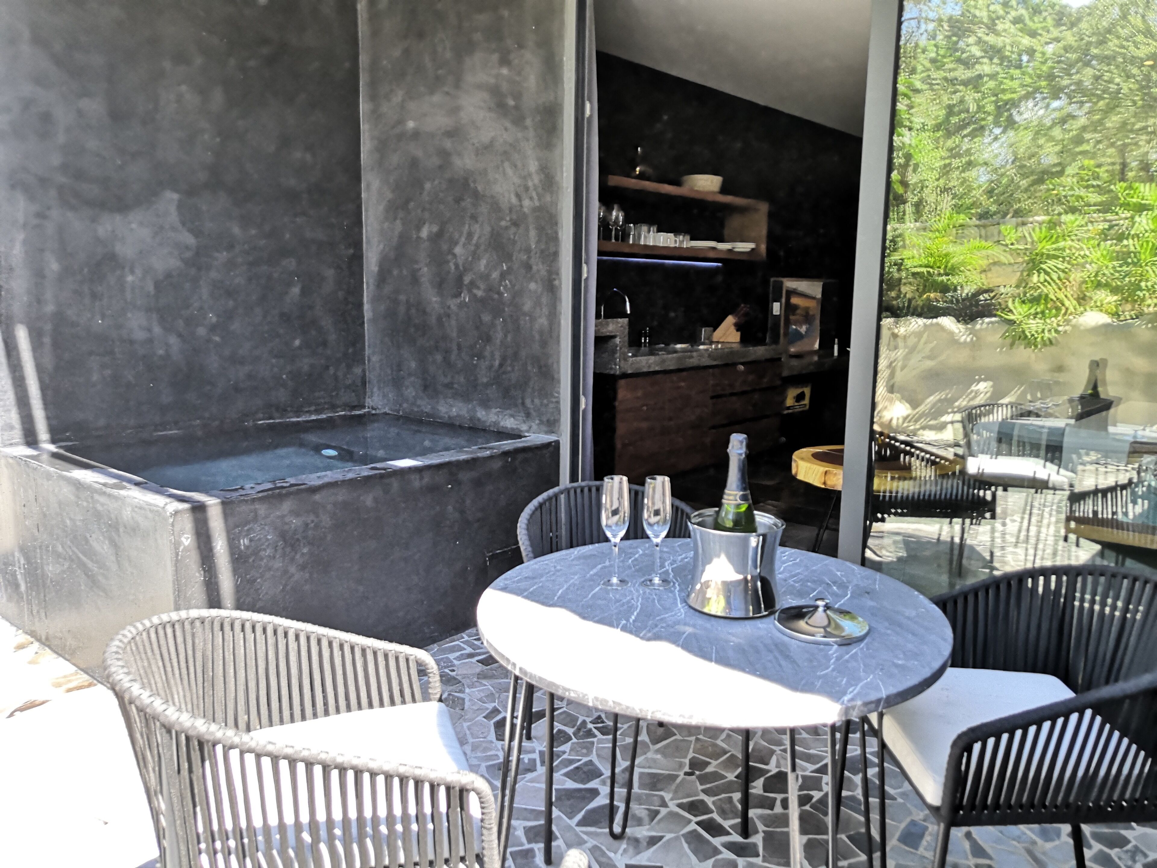 Une terrasse extérieure avec un sol carrelé en pierre, une table ronde en marbre, des chaises en osier et un seau en métal contenant une bouteille et des verres.