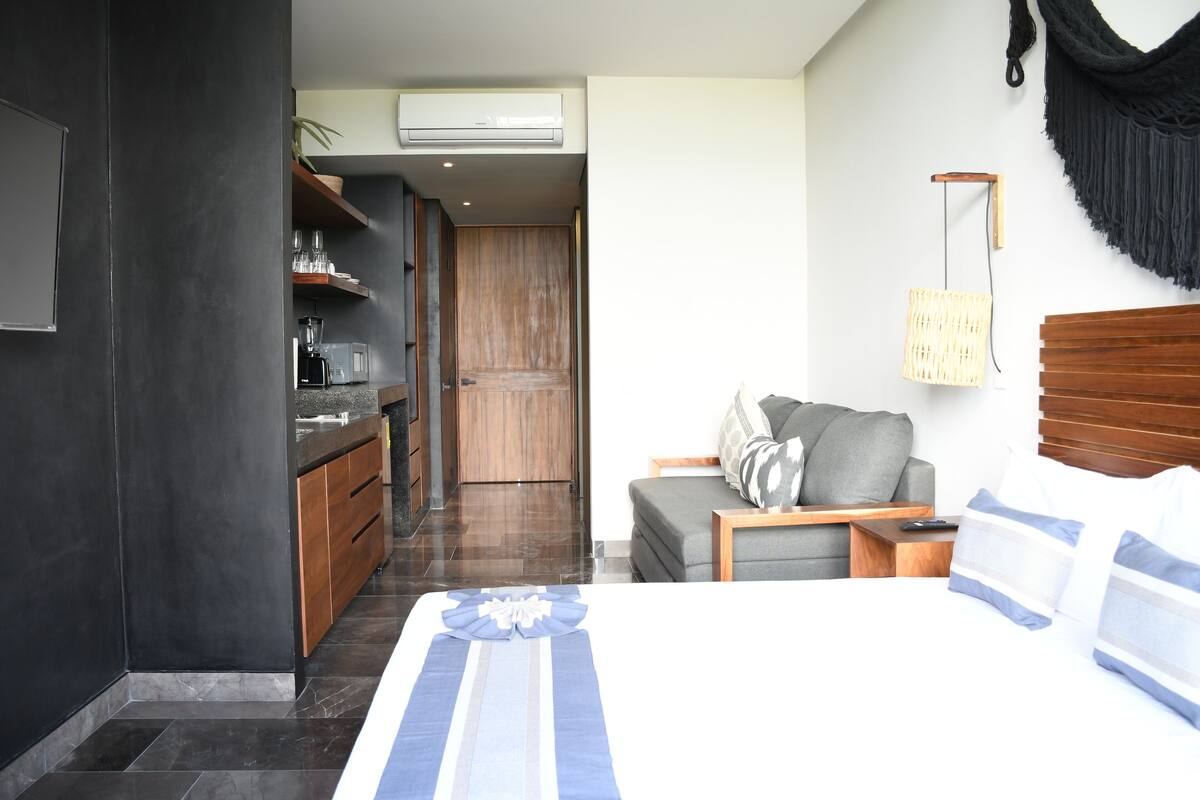A modern hotel room with a large bed, a sofa, a small table, and a kitchen area with a coffee machine.