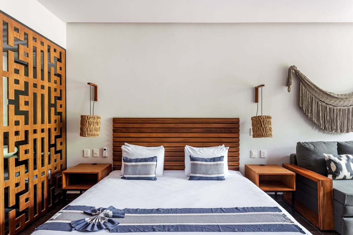 A modern hotel room with a wooden headboard, two bedside tables with lamps, a grey sofa, and a decorative wall panel.