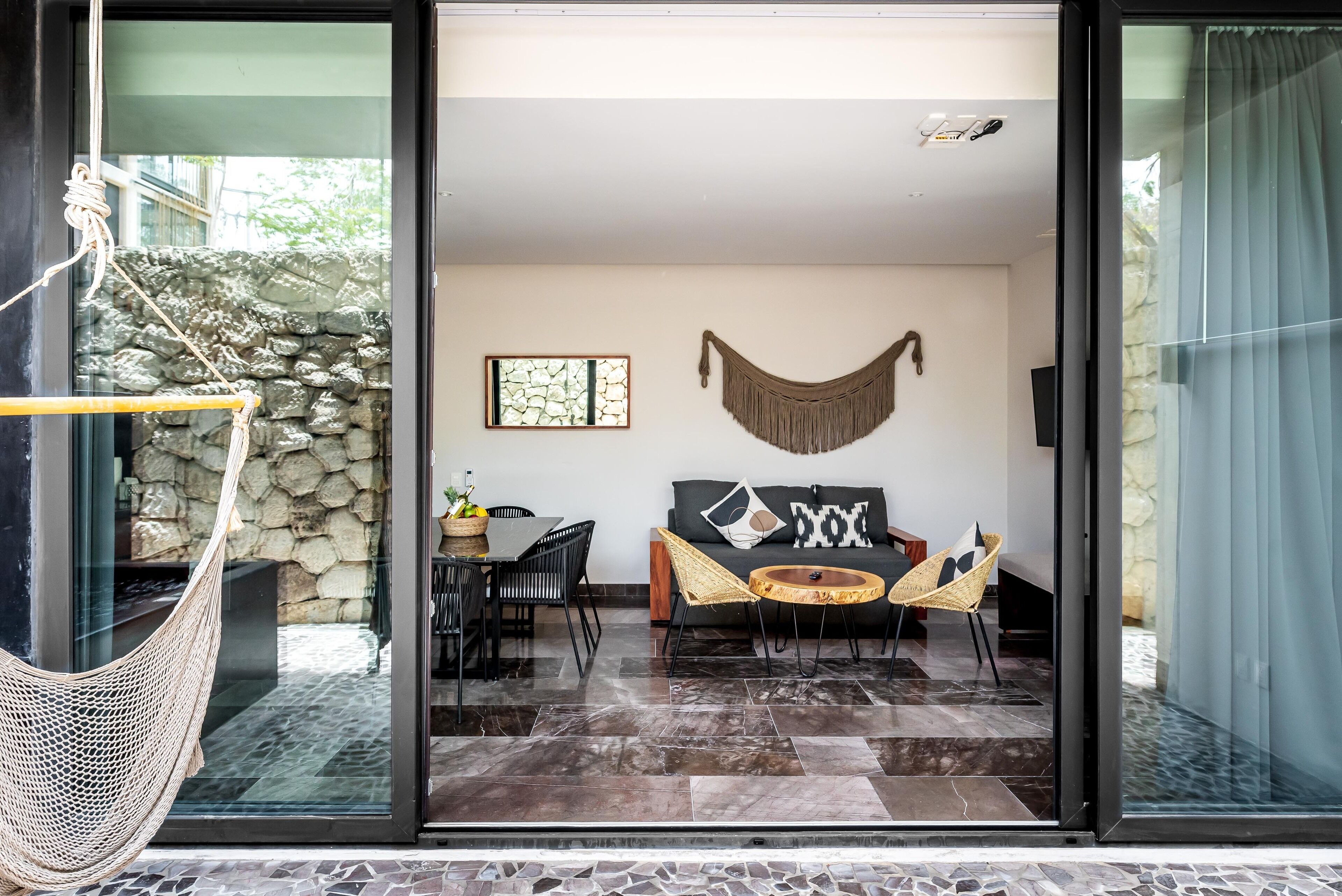 Un intérieur moderne avec un mur en pierre, un hamac, un canapé avec des coussins, une table basse et un coin repas avec des chaises.