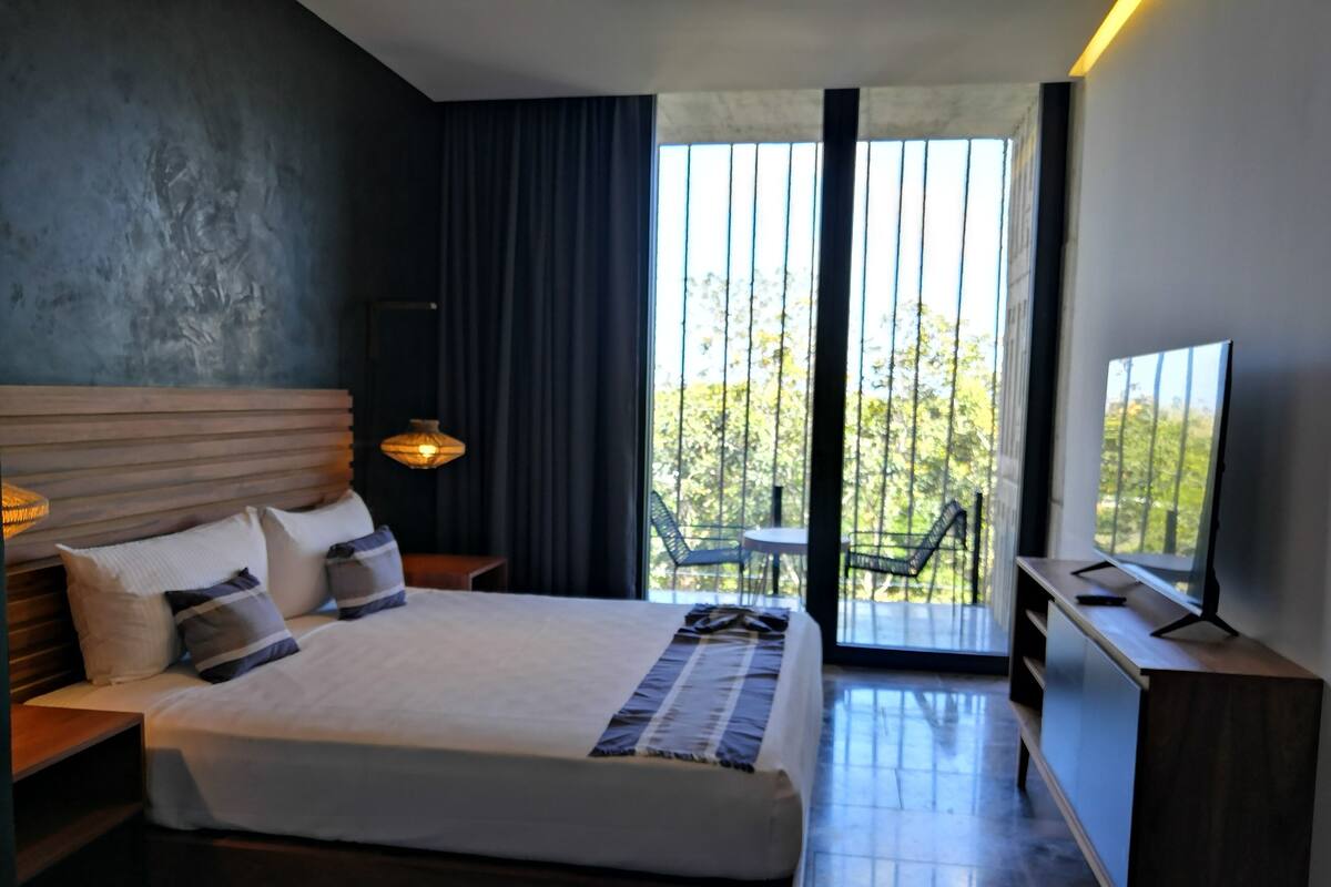 A modern hotel room with a large bed, a TV, and a balcony with outdoor seating.