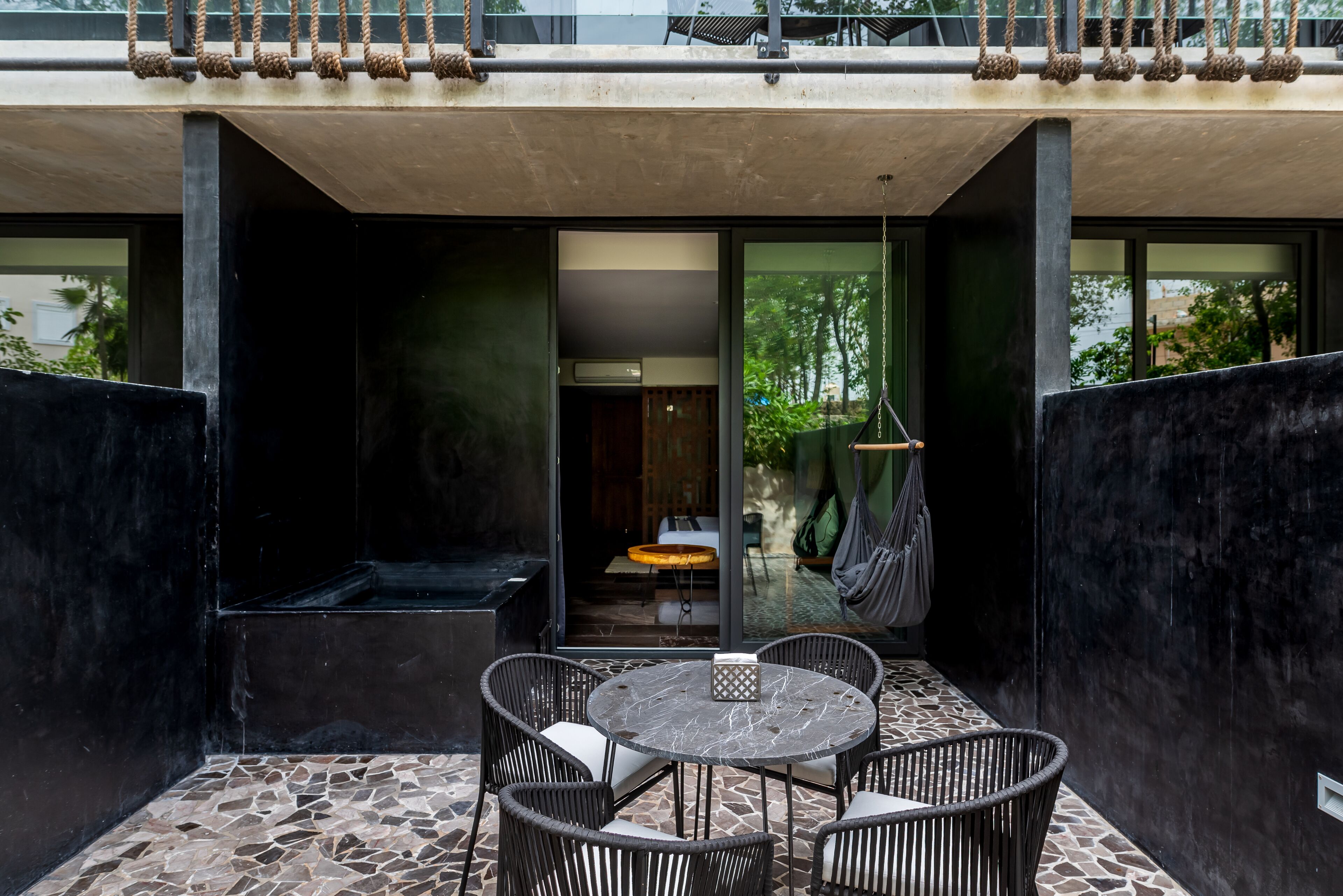 En moderne udendørs terrasse med et rundt bord, fletstole, en hængekøje og udsigt til indretningen gennem glasdøre.